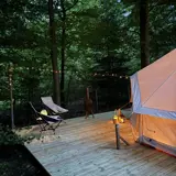 Glampingtelt på træterrasse foran skov i aftenlys, oplyst af lyskæder og lanterner der skaber en hyggelig stemning.